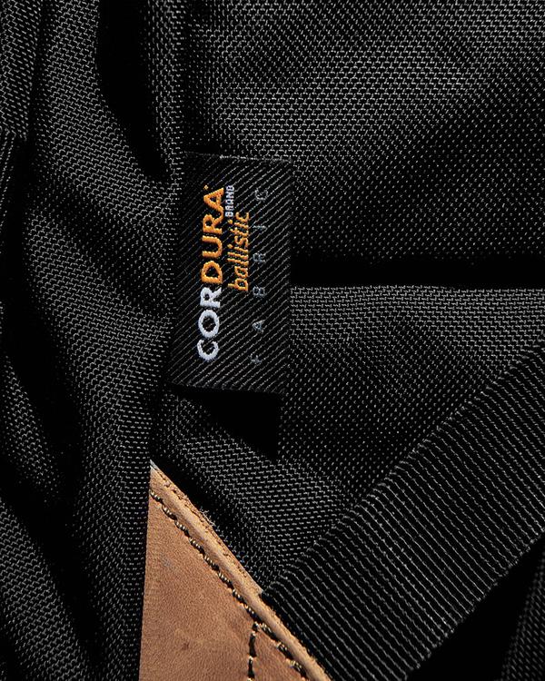 Close-up of a black fabric label on the FXD WBP-3 Work Backpack, showcasing "CORDURA Ballistic fabric" in orange and white. The label is sewn onto black material with a partially visible brown leather base.
