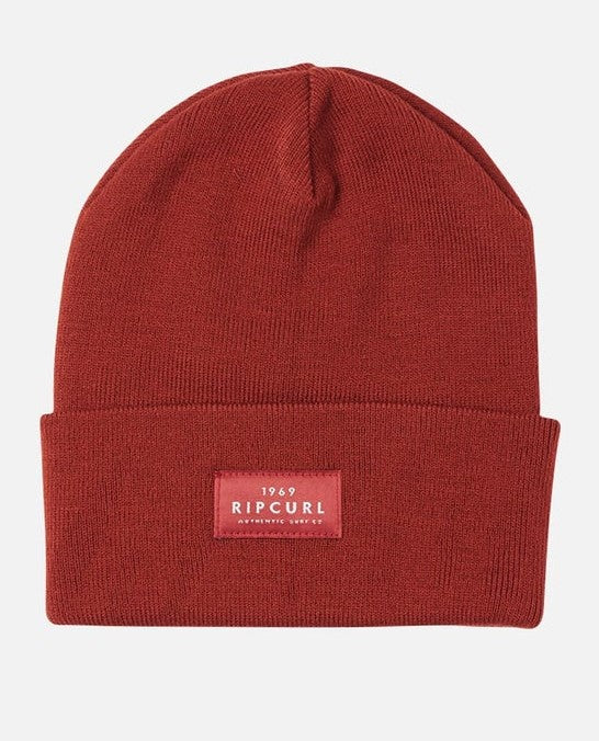 The Rip Curl Premium Surf Beanie is a red rib knit beanie with a folded brim, featuring a rectangular label on the front that reads "1969 RIP CURL Authentic Wet Suits." The beanie is displayed against a white background.
