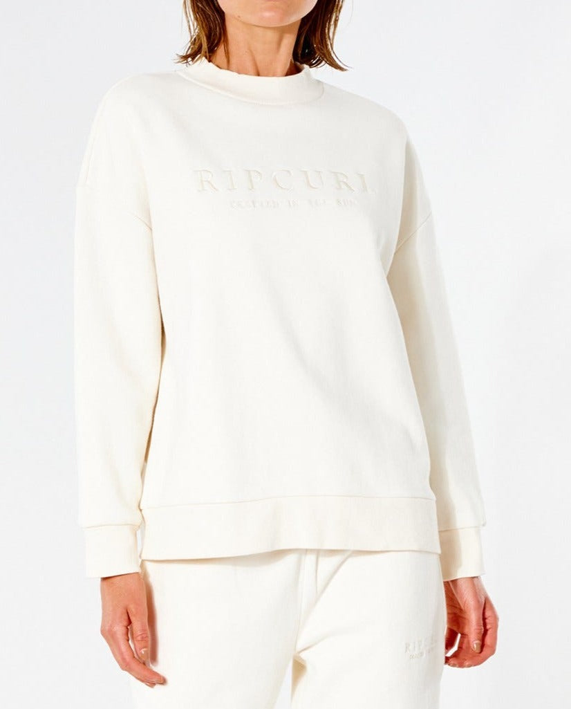 A person wears a cream-colored Rip Curl Premium Surf Crew jumper with "RIPCURL" on the front, paired with matching cream pants. The casual jumper features a soft fleece lining, set against a plain white background, and is cropped at the neck and mid-thigh.