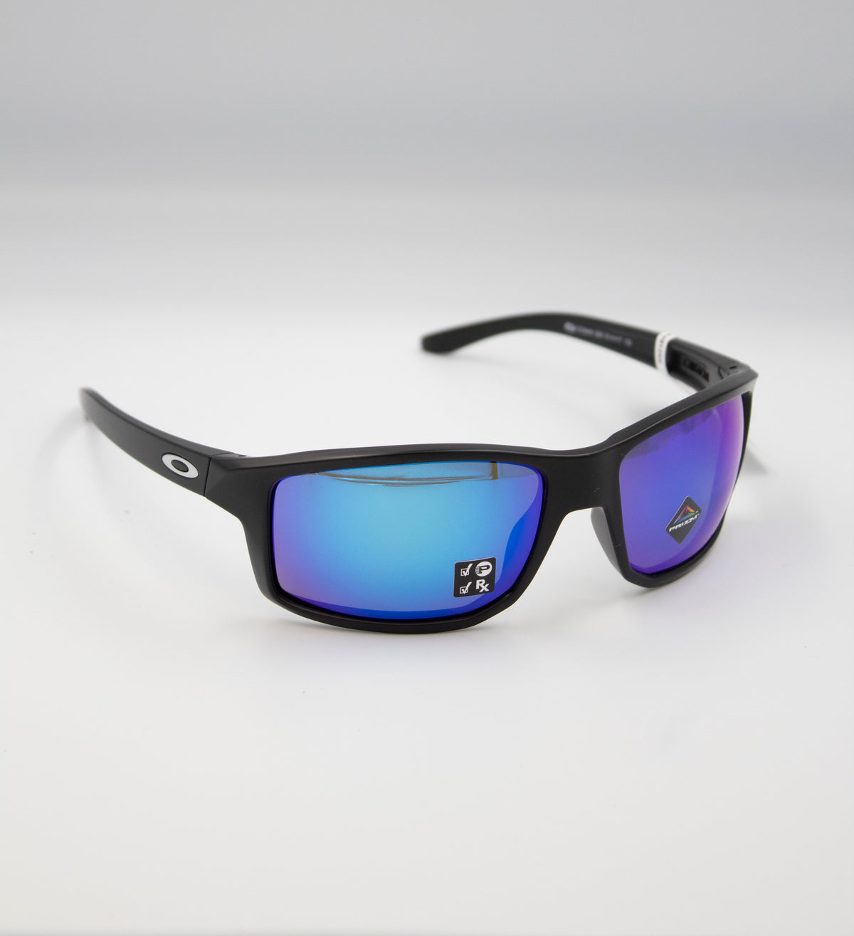 A pair of Oakley Gibston Polarized Matte Black Sunglasses rests on a white surface. These black-framed sunglasses include blue mirrored Prizm lenses, a small logo on the temple, and a sticker on one lens. Their sleek and modern design makes them perfect for both casual and sporty looks.