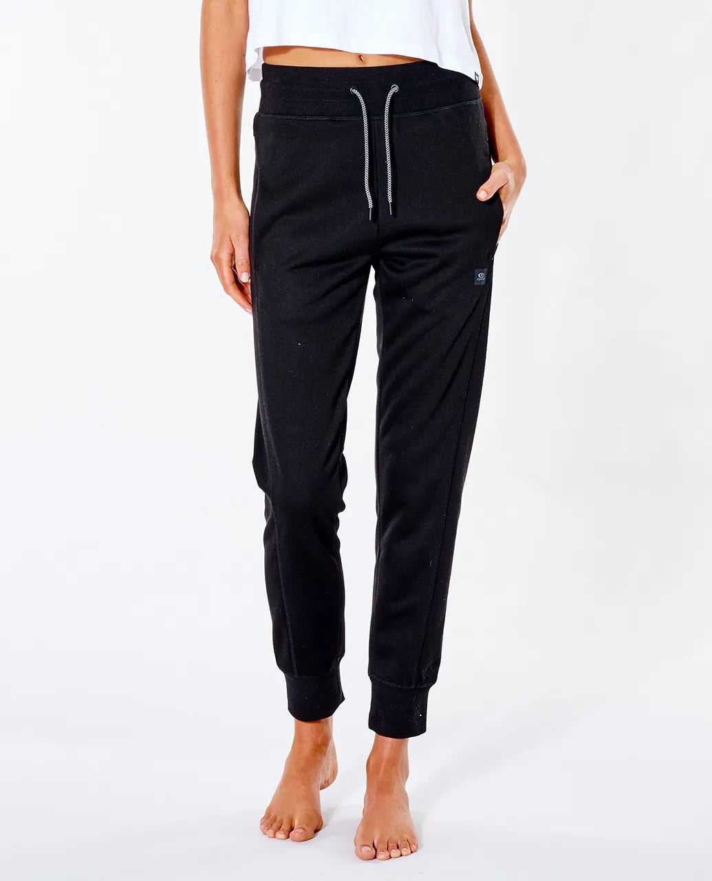 A person standing barefoot on a white background is wearing Rip Curl Anti Series Flux II Trackpants with a drawstring waist and pockets. They pair the slim-fit, black jogger pants, which have cuffed ankles and are made from durable water-resistant fabric, with a cropped white top. Their right hand rests in one of the jogger's pockets.
