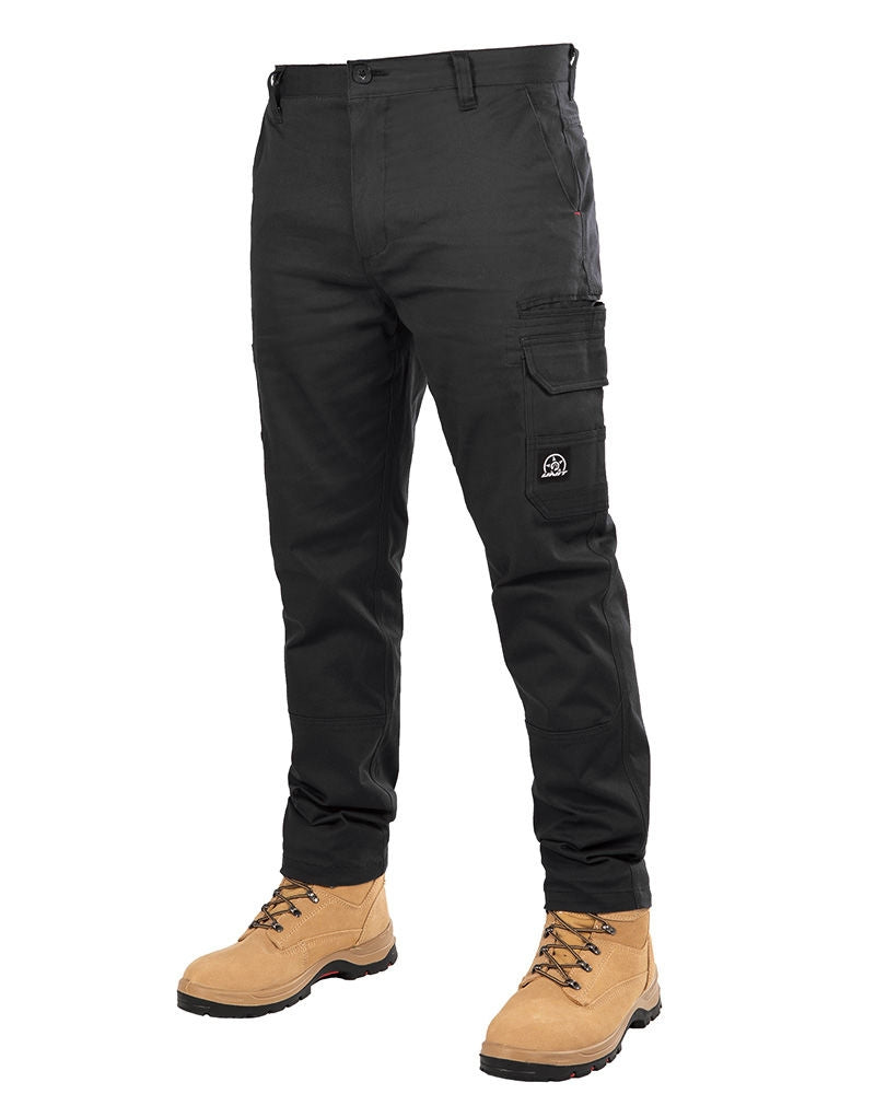 A person is wearing the UNIT Demolition Stretch Cargo Pant in durable black, featuring multiple pockets and a small white logo on the thigh pocket, paired with tan work boots. The pants offer UPF 50+ protection. The image focuses on the lower half of the person, showcasing the leg area and footwear in detail.