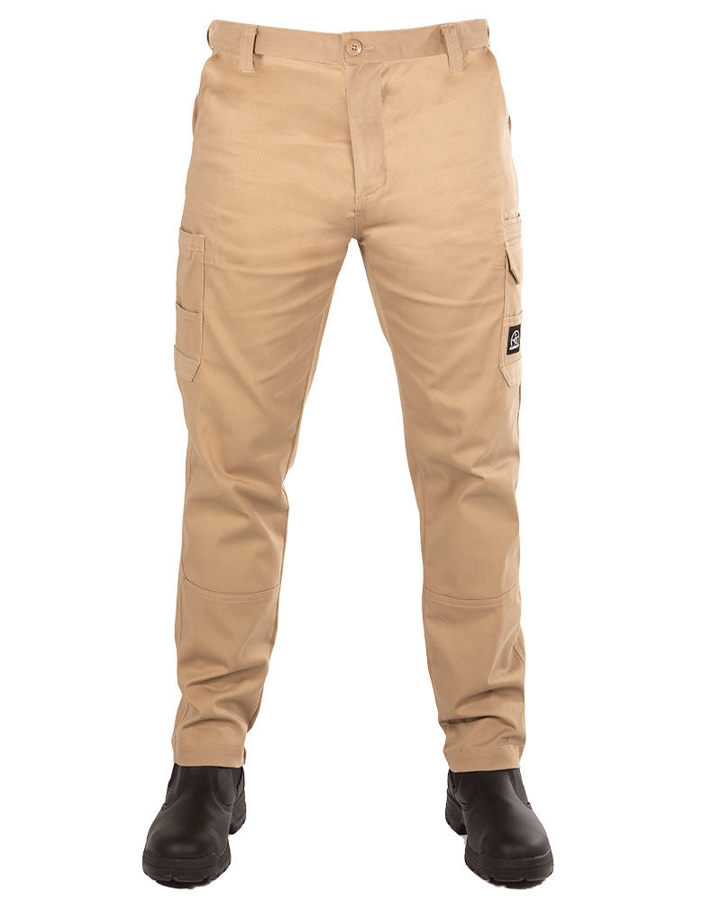 A person is wearing the UNIT Demolition Stretch Cargo Pant, a pair of durable beige work pants offering UPF 50+ protection and multiple cargo-style pockets. The pants feature a button and zipper closure at the waist and are paired with black work boots. The individual stands with their legs slightly apart.