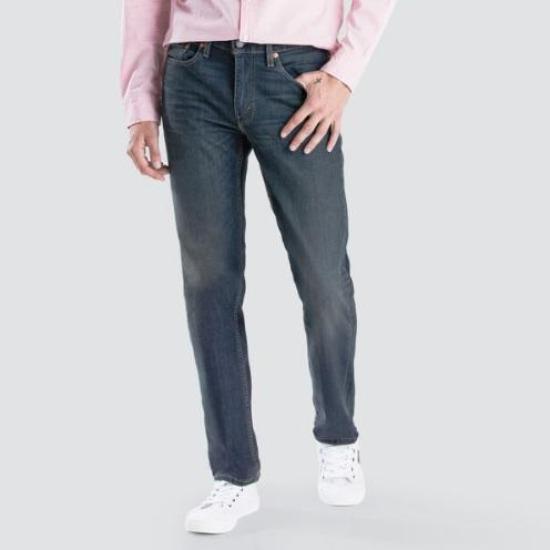 A person is standing against a gray background wearing a light pink long-sleeve shirt, stylish and comfortable Levis 514 Straight Jean, and white sneakers. Only the torso and legs are visible. One hand is placed in a front pocket, and the other hand is hanging by the side.