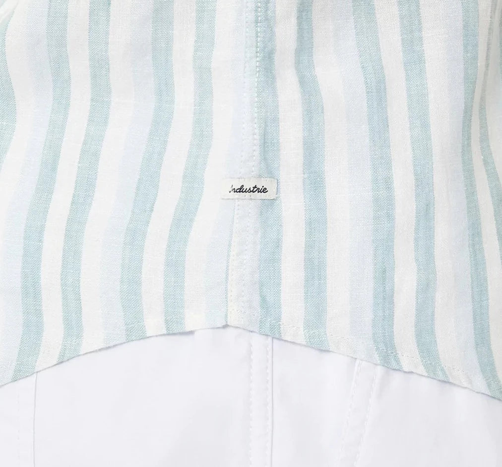 Close-up of Industrie The Linenman Shirt, featuring vertical light blue and white stripes with a button-down collar. A black "industrie" script fabric label is at the seam, while the lower part maintains a plain white finish in this relaxed fit shirt.