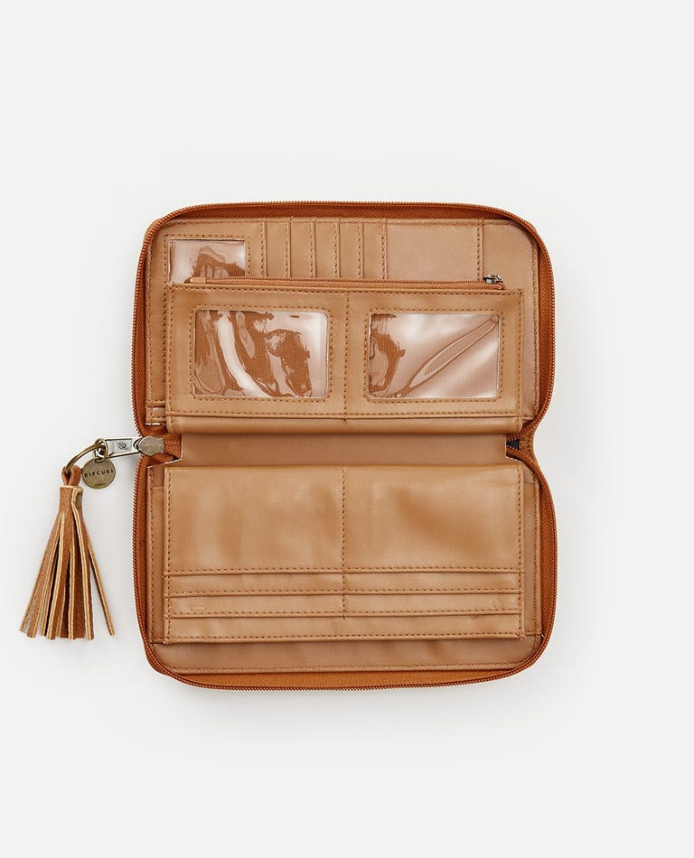 The Rip Curl Surf Gypsy Oversized tan leather wallet is open to reveal several compartments, including card slots, a zippered pocket, and transparent ID windows. An optional removable tassel keyring is attached to the zipper pull on the left side of the wallet. The background is white.
