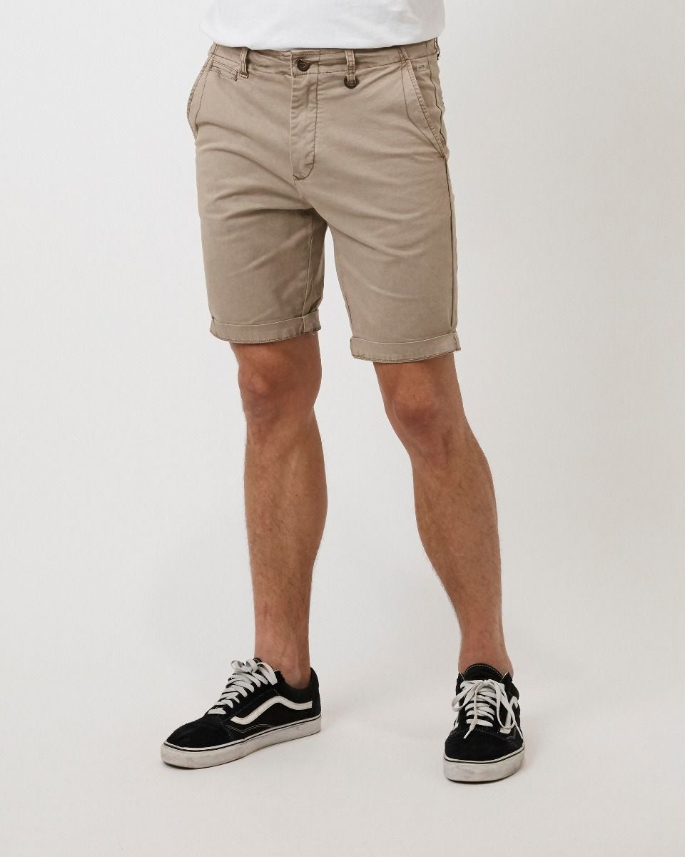 A person is standing against a plain white background. They are wearing beige knee-length shorts from Industrie The Rinse Short collection, black and white sneakers, and a comfortable white shirt (partially visible). The person's hands are not visible in the frame.