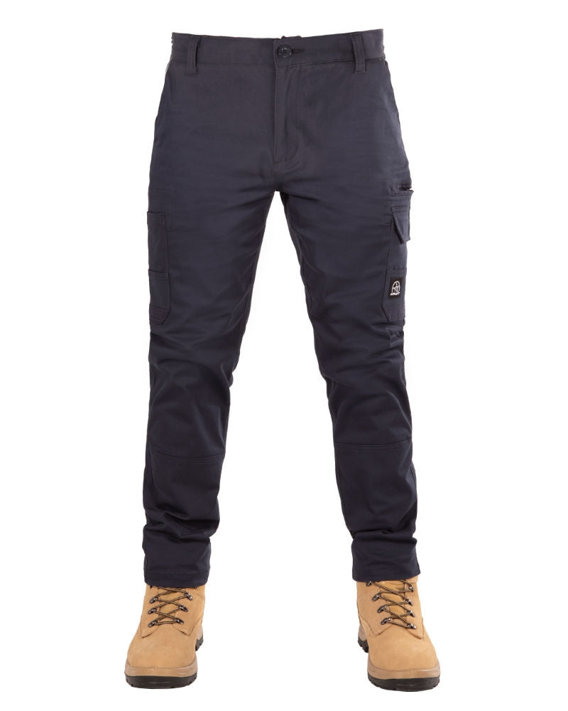 A person is wearing durable UNIT Demolition Stretch Cargo Pants in navy blue with multiple pockets and tan work boots with light brown laces. The slim-fit pants, featuring UPF 50+ protection, create a practical yet stylish look. The person's hands are not visible in the white background image.