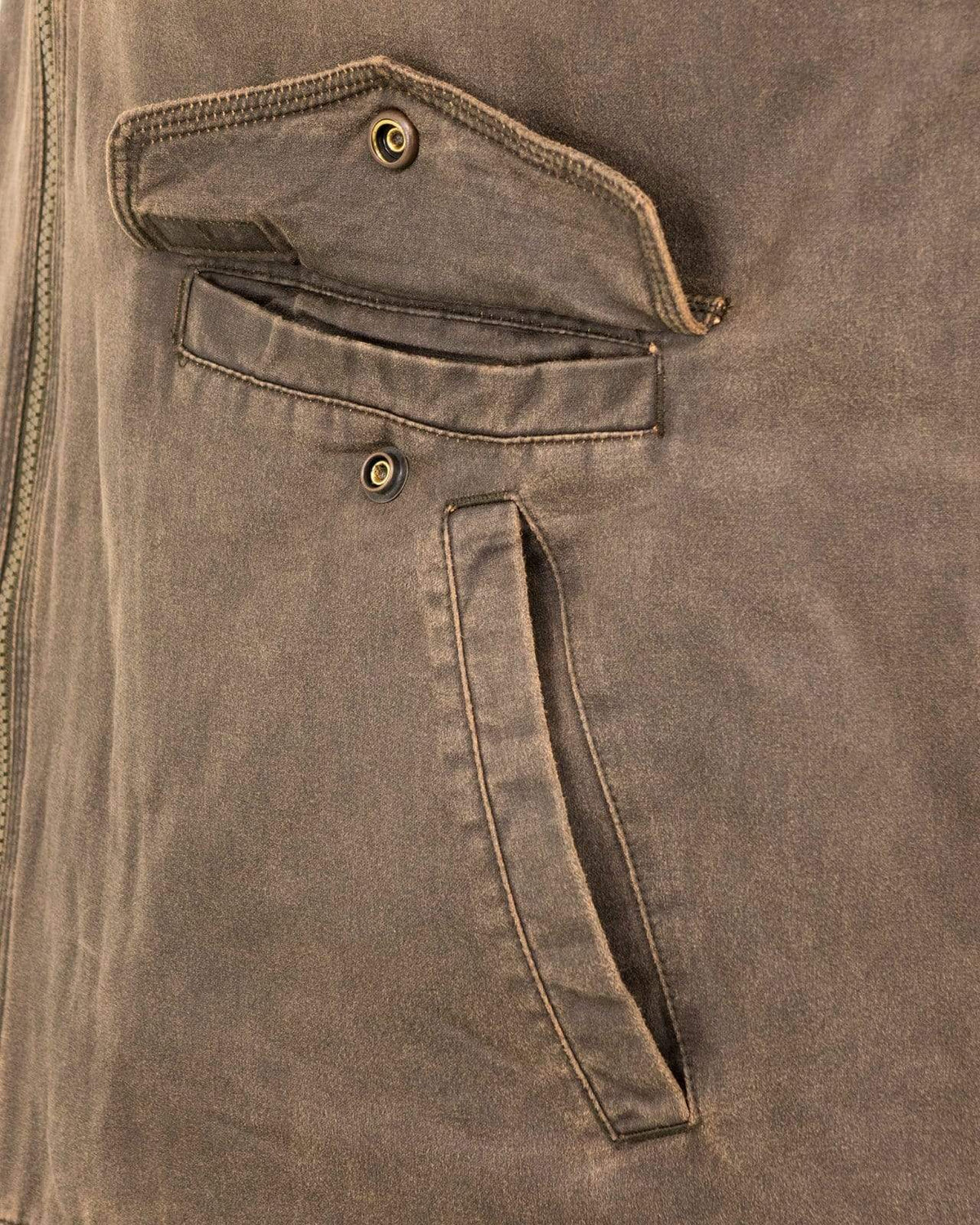 Close-up of the Outback Landsman Vest's pocket area, showcasing two pockets with button details. The top pocket features a flap with one button and is partially open, revealing the interior. The bottom pocket is a simple slit pocket with visible stitching. The machine washable fabric has a weathered look.
