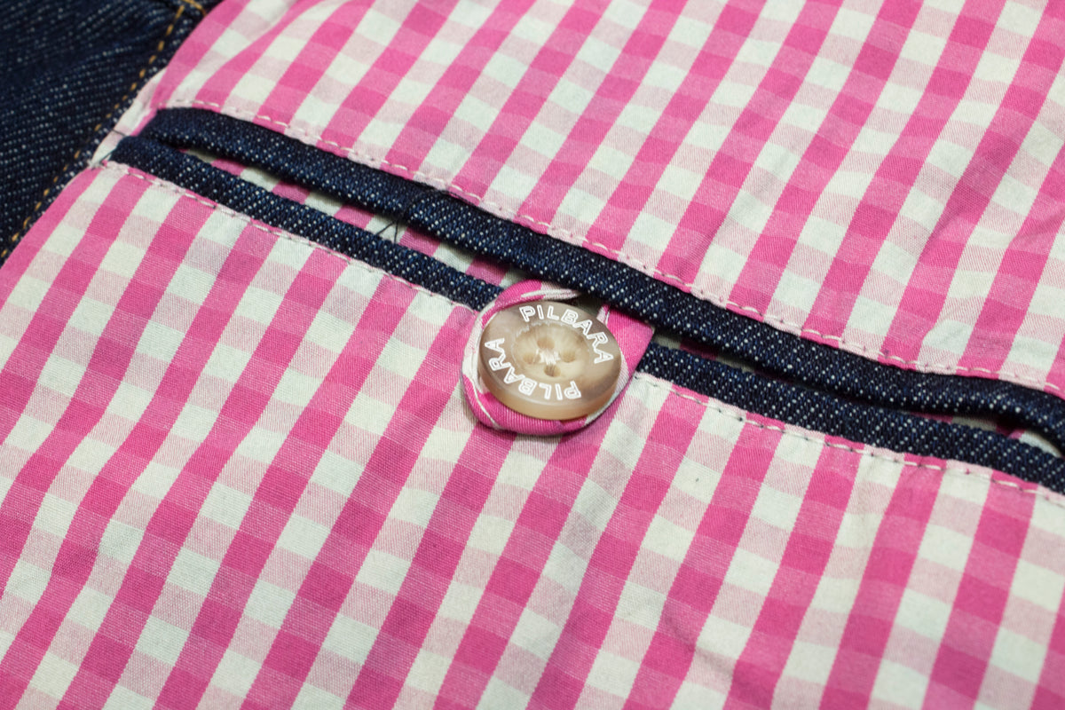 Close-up of a pink and white gingham from the Pilbara Ladies Denim Jacket featuring a pink button with "PILGRIM" repeatedly. The 100% cotton fabric is detailed with contrasting dark blue denim trim.