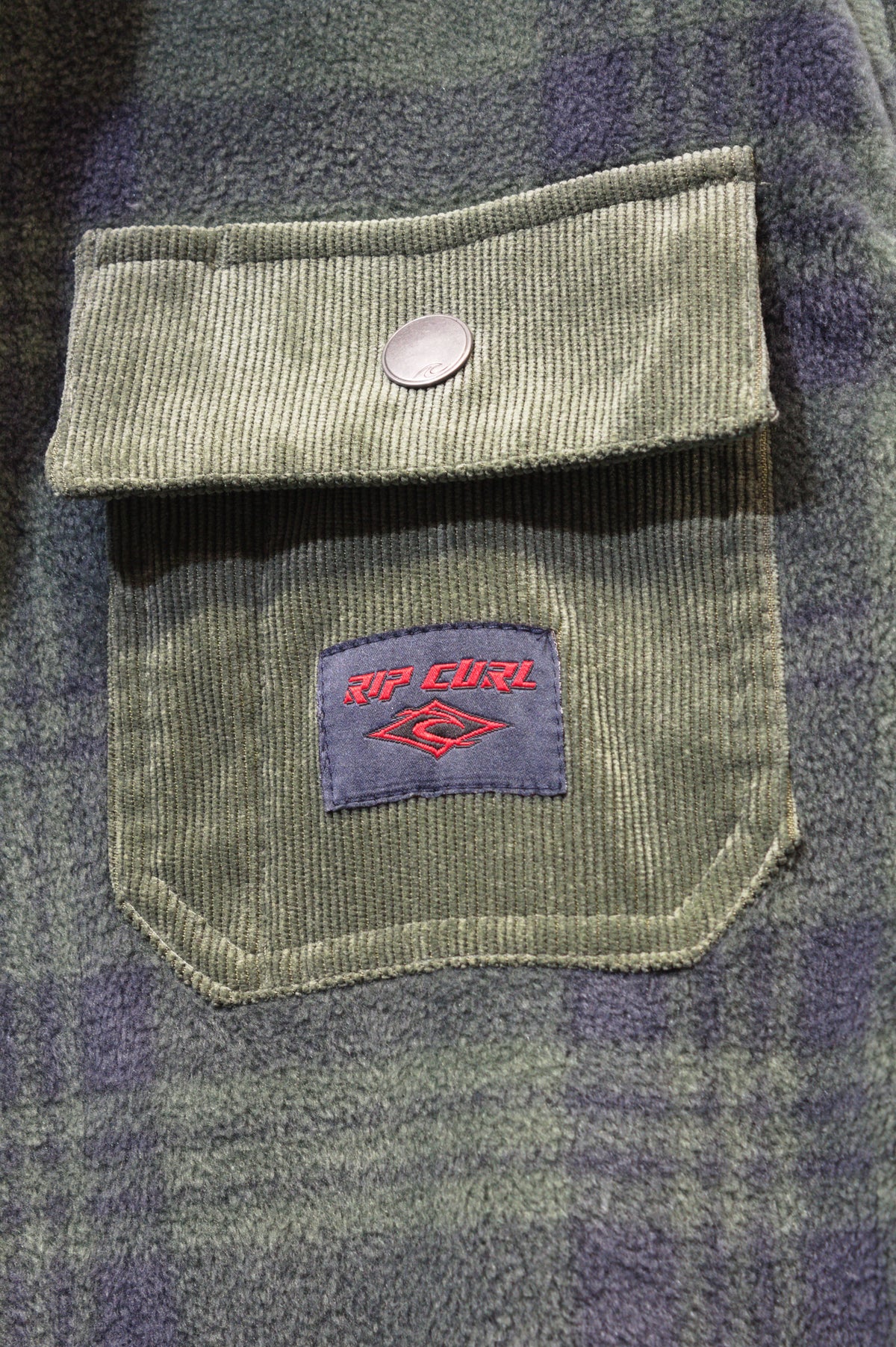 Close-up of the Rip Curl Polar Fleece Party Pack Shirt in green plaid, featuring a corduroy texture and a buttoned flap pocket adorned with a rectangular red and blue "Rip Curl" logo patch, highlighting its unique design.