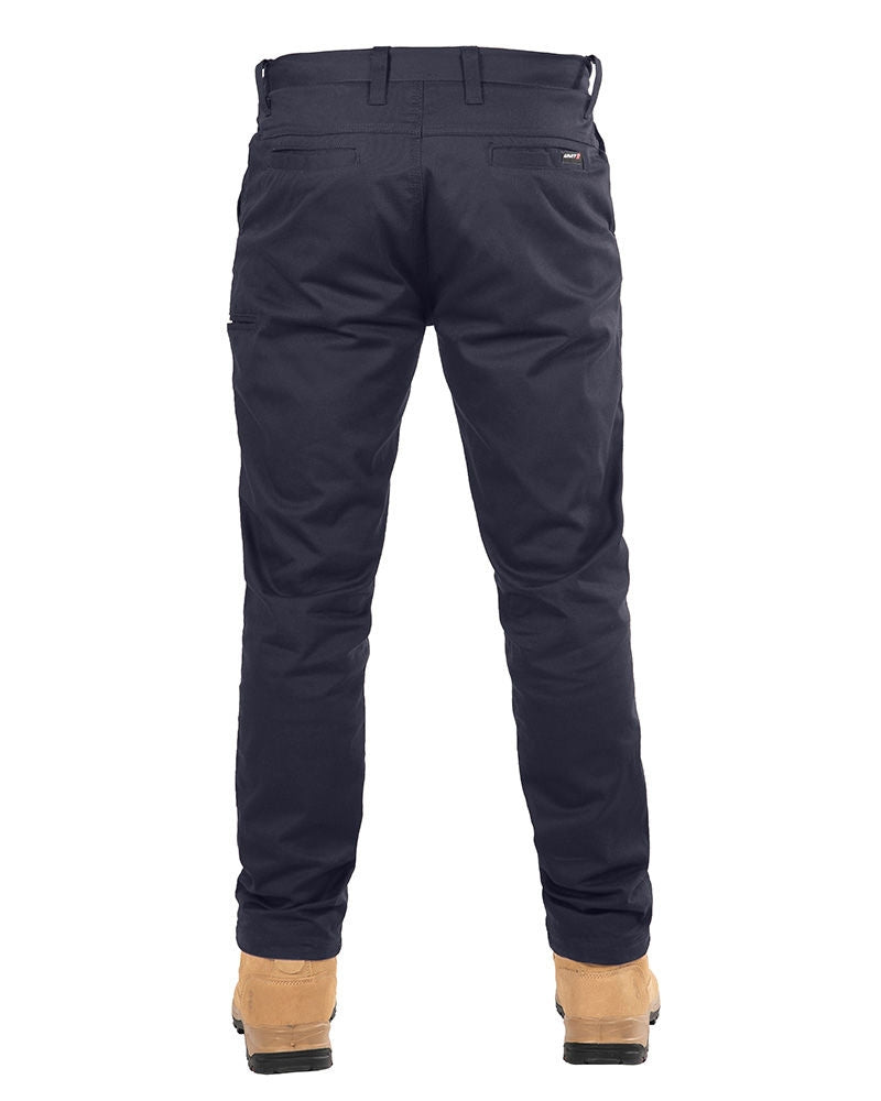 A rear view of a person wearing navy blue UNIT Ignition Stretch Pants and tan work boots, standing with legs slightly apart. The pants, made from stretch fabric, feature belt loops, back pockets, and a stitched logo patch on the right rear pocket. The person is standing against a plain white background.