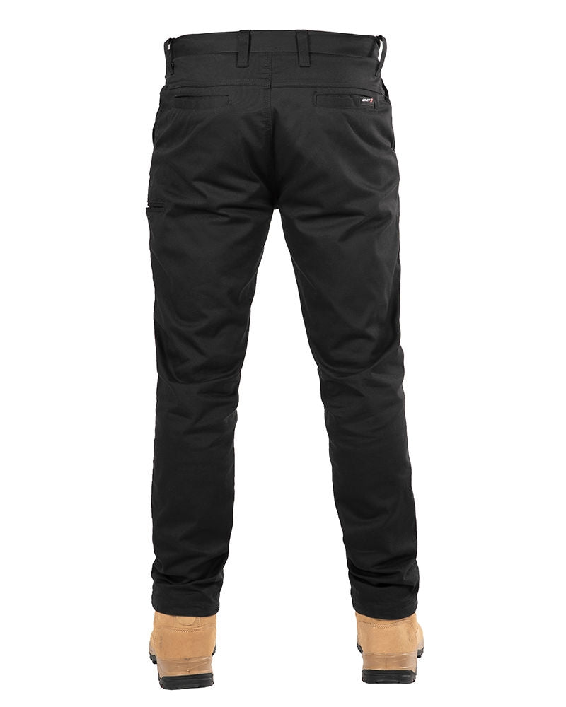 A person wearing UNIT Ignition Stretch Pants and tan work boots is shown from the back. The pants, designed as durable workwear, feature belt loops, welt pockets, and appear to be crafted from thick stretch fabric. Only the lower part of the person's body is visible against a plain white background.