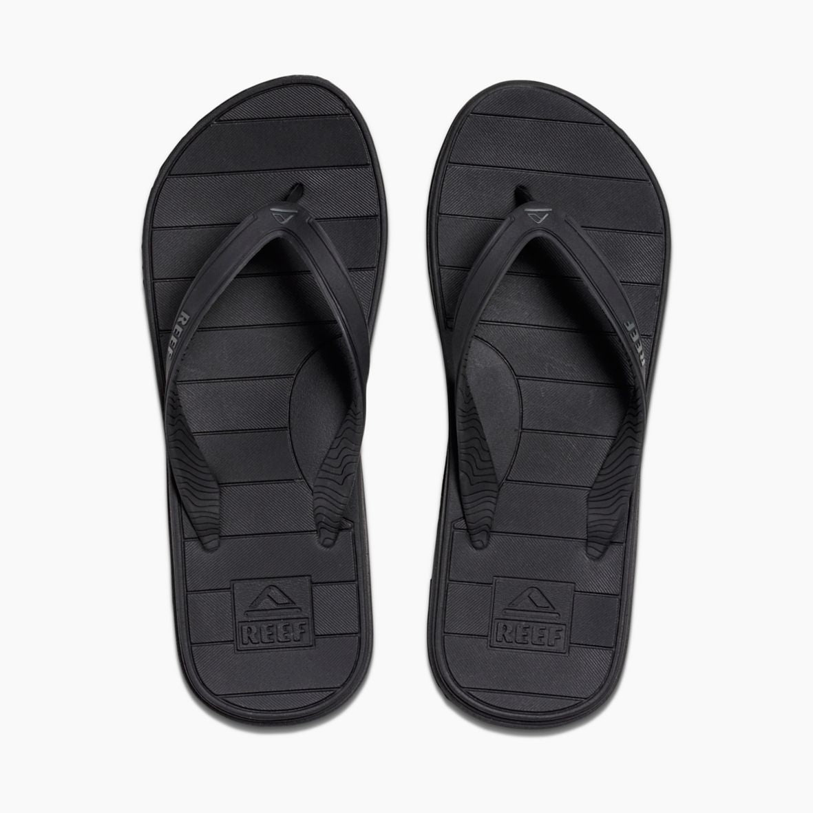 A pair of REEF Switchfoot LX flip-flops in black features a simple striped pattern on the rubber sponge footbed. The brand name "Reef" is embossed on the heel area of each sandal, with subtle text and logo detailing on the straps. These flip-flops, designed with arch support, are displayed against a plain white background.