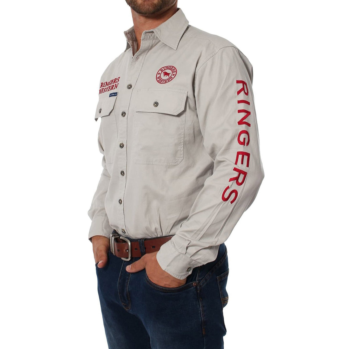 A man stands against a white background in a Ringers Western Hawkeye Embroidered Shirt, featuring "RINGERS" on the sleeve and chest patches, made from 100% cotton. He pairs it with dark jeans and a brown belt, resting his hands casually in his pockets.
