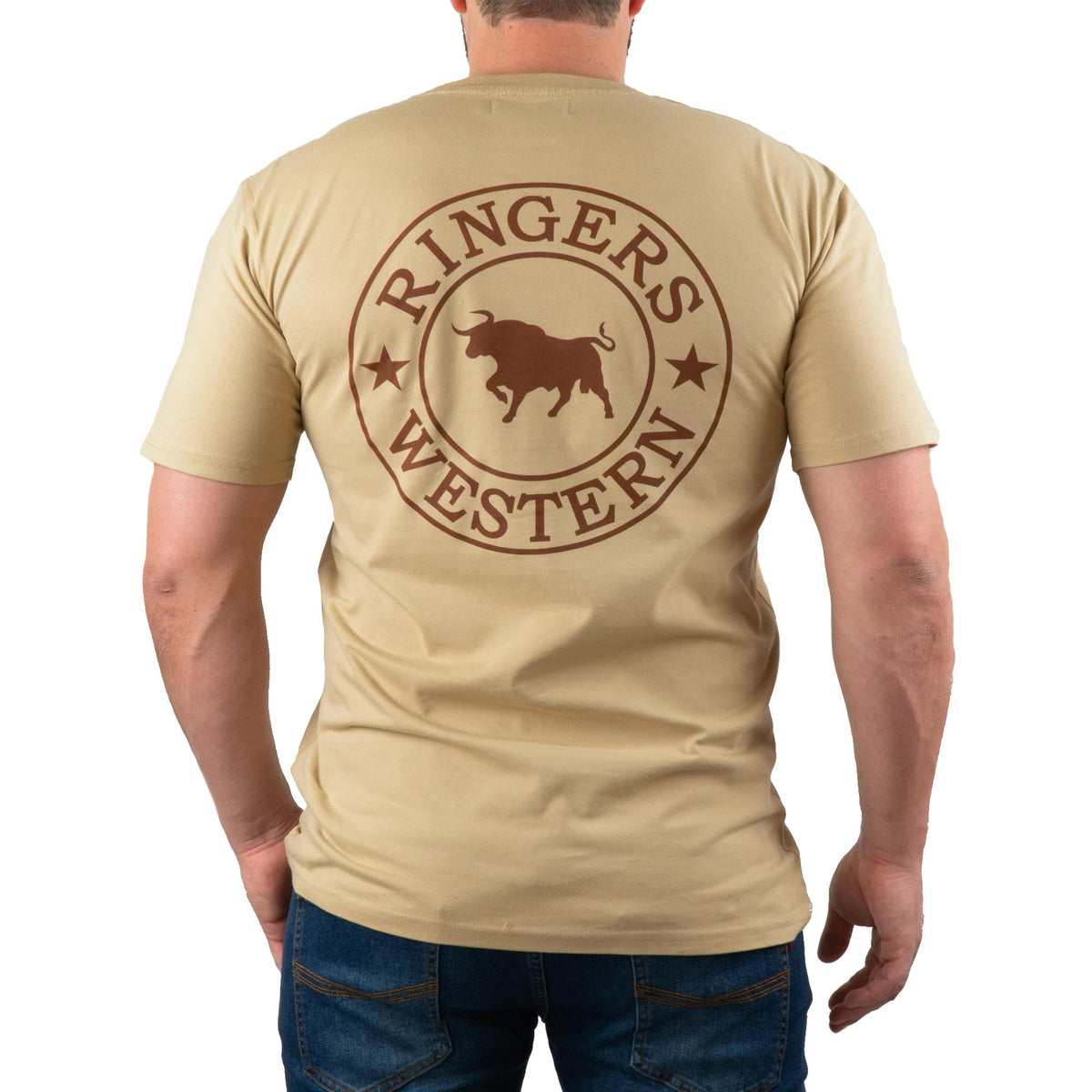 A man is wearing the Ringers Western Signature Bull Classic T-Shirt in beige, which features a comfortable crew neck design. On the back, there's a circular emblem with a bull in the center, surrounded by the text "RINGERS WESTERN" and two stars. The man is facing away from the camera and is also dressed in blue jeans.