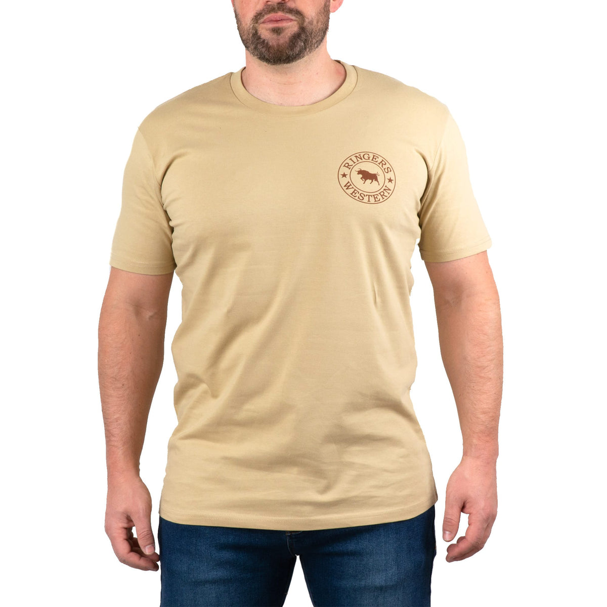 A man wearing a comfortable beige Ringers Western Signature Bull Classic T-Shirt, which features a brown circular emblem with a small horse design and text on the left chest. The crew neck t-shirt is paired with blue jeans, and he is standing against a plain white background.