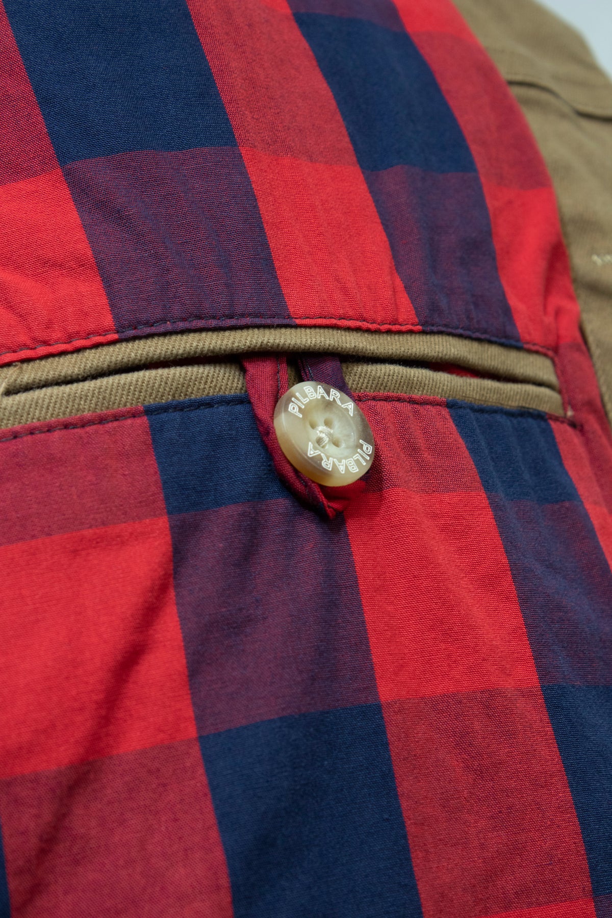 Close-up of a classic fit plaid fabric in alternating red and blue squares with a beige horizontal zipper pocket. The pocket flap, part of this Ritemate Pilbara Cotton Canvas Jacket, is fastened with a brown button that has the word "Pulabo" engraved on it.