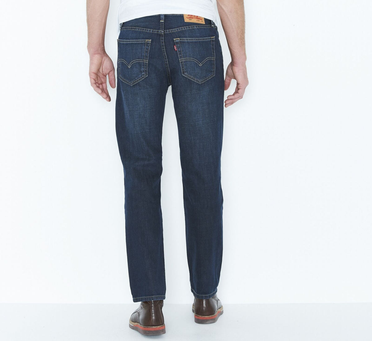 A person wearing a pair of dark blue Levis 516 Straight Jeans and a white T-shirt is standing with their back to the camera. The 100% cotton jeans feature visible stitching on the pockets and a brown leather patch on the back. The person is also sporting brown shoes with red soles.