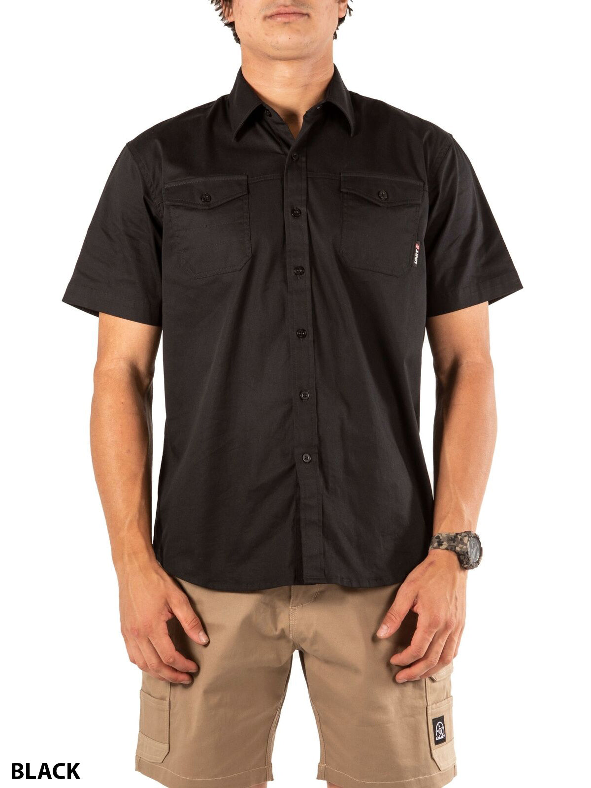 A person wears the UNIT Craftman Stretch Short Sleeve Shirt in black with two chest pockets and tan cargo shorts. The stretch-fit shirt offers comfort as they stand facing forward, hands by their sides.