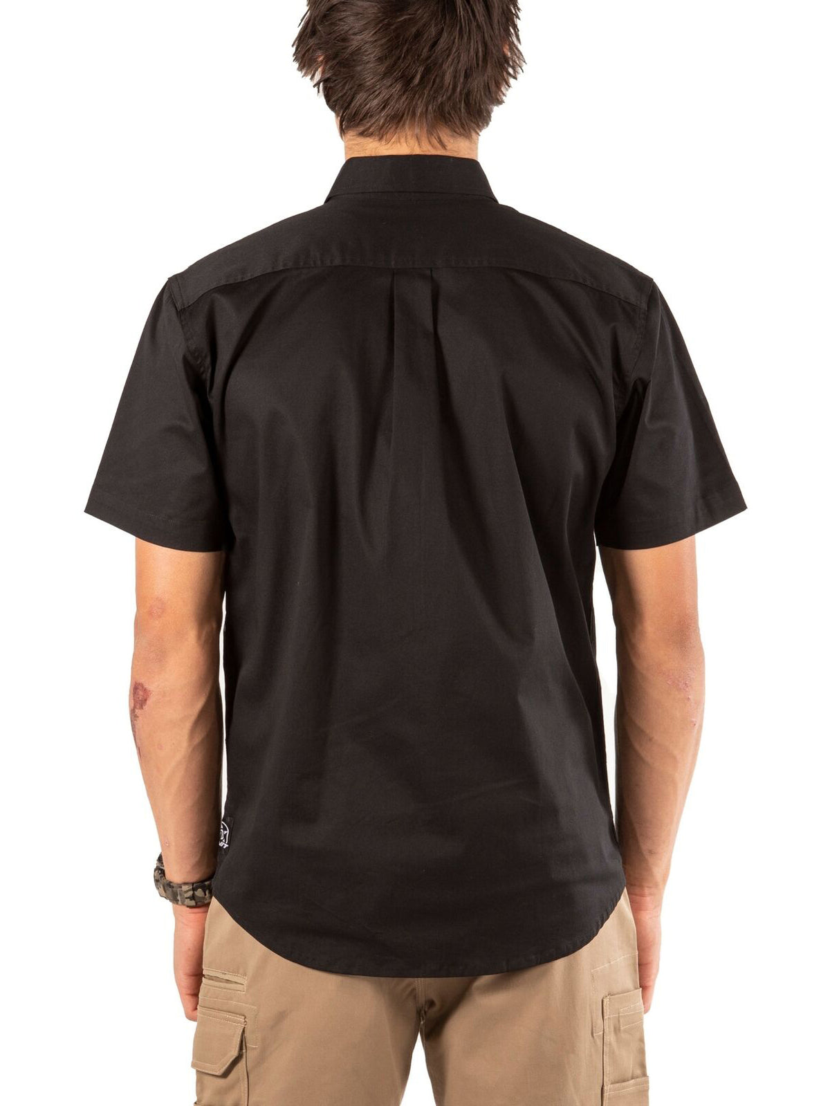 A man with short brown hair stands facing away, wearing the UNIT Craftman Stretch Short Sleeve Shirt in black and khaki cargo shorts, plus a watch on his left wrist. The background is plain white.