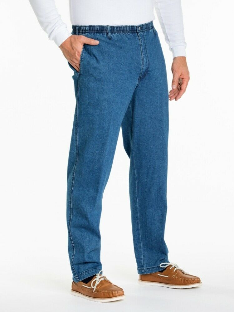 A person is wearing blue Breakaway Ipswich Denim Jeans with an elastic waistband and a mid-rise design, a white long-sleeve shirt, and light brown moccasin-style shoes. The person's left hand is in the pocket of the jeans, and the background is white.