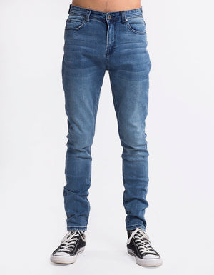 Against a white background, someone is wearing Silent Theory Shadow Jean in blue denim, featuring a cropped skinny fit with button closure and visible stitching, paired with black high-top sneakers with white laces and toe caps.