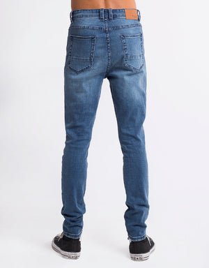 A person is seen from behind wearing Silent Theory Shadow Jean, cropped skinny fit blue jeans with visible stitching, paired with black and white sneakers. The jeans have two back pockets and a leather patch on the waistband, against a plain white background.