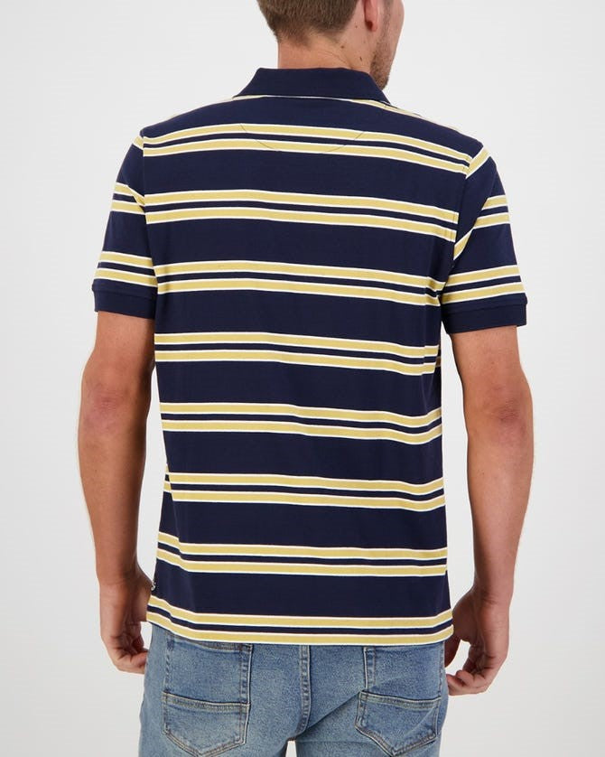 A person wearing a Swanndri Lincoln Stripe Polo, featuring navy blue with yellow and white horizontal stripes, and blue jeans is shown from the back. The 100% cotton casual polo shirt complements their short hair as they stand against a plain, light-colored background.