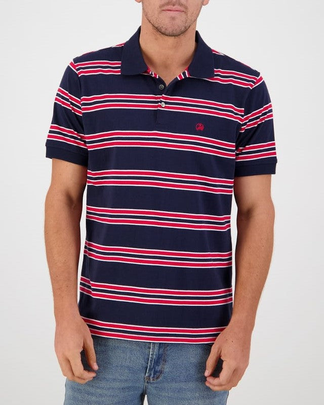 A person is wearing a Swanndri Lincoln Stripe Polo in navy blue with red, white, and blue horizontal stripes. The 100% cotton shirt features a small red logo on the left chest. They are also wearing blue jeans and posing against a plain white background. The person's face is not visible.