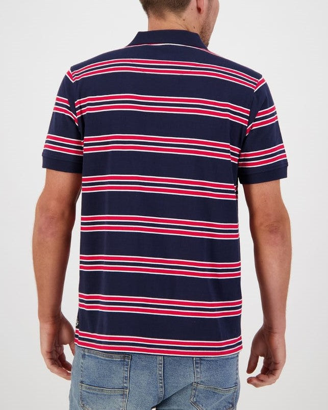 A person with short hair is seen from the back wearing the Swanndri Lincoln Stripe Polo, featuring casual navy blue fabric with red and white horizontal stripes made of 100% cotton. They are also wearing blue jeans against a plain white background.