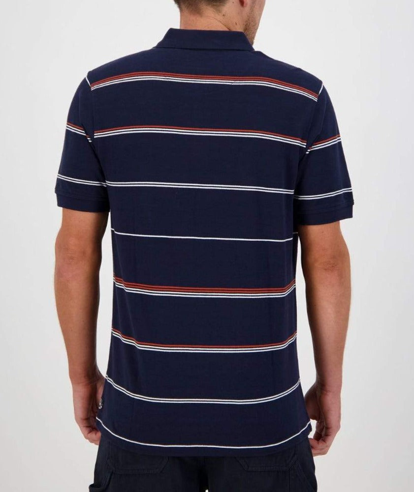 A person is wearing a Swanndri Concord Stripe Polo, characterized by its navy blue short sleeves and horizontal white and red stripes. Featuring the distinct Swanndri logo, they stand with their back to the camera, arms slightly bent at the elbows. The background is plain and light-colored.
