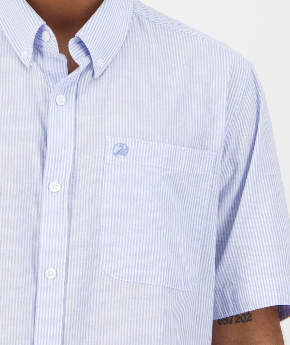 A person with a small tattoo on their arm wears the Swanndri Hinsdale Shirt, featuring light blue and white stripes, a button-down collar, chest pocket with an embroidered logo. This 100% cotton shirt offers a comfortable, classic fit with short sleeves.