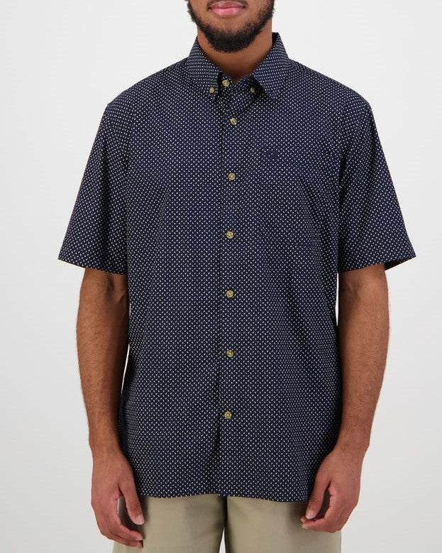 A person models against a plain backdrop in the Swanndri Putney Shirt, a tailored-fit navy short-sleeve button-up featuring a subtle white pattern. This 100% cotton shirt with gold buttons complements their beige pants and beard effortlessly.