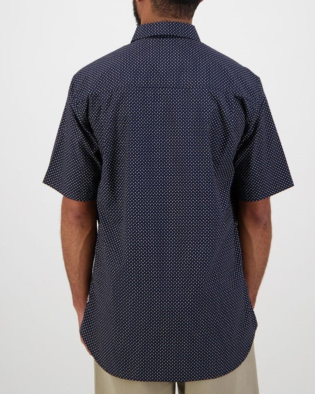 The individual is shown from behind wearing a tailored-fit Swanndri Putney Shirt, a dark short-sleeve shirt with small white polka dots made from 100% cotton. Paired with beige pants, this ensemble stands out against the plain, neutral background.