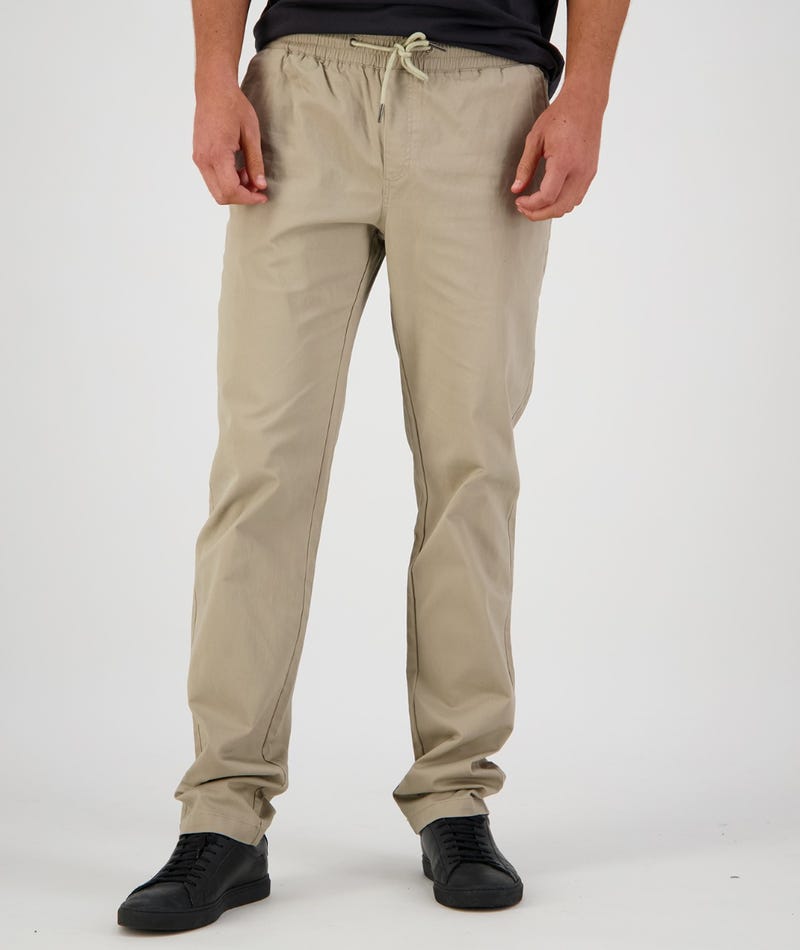 A person stands against a plain background, modeling Swanndri Karaka Pant in beige with an elastic waist and black sneakers. The white drawstrings are tied in a bow at the waist. The person is wearing a dark top that is partially visible in the frame.