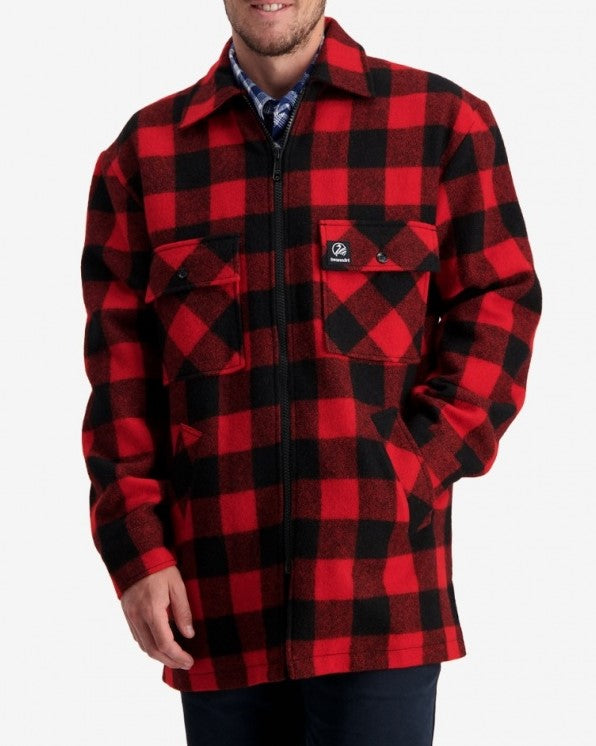 A person is wearing the Swanndri Longford Wool Shacket, a red and black plaid checkered overshirt made of 100% wool, featuring two front chest pockets and a zipper closure. The individual is standing against a plain white background, with their left hand in the jacket pocket.