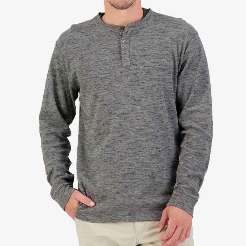 A person is shown from the shoulders down, wearing a Swanndri Oxford Henley shirt in grey with long sleeves, featuring three buttons at the neckline and waffle knit detailing. Made from 100% cotton, it pairs perfectly with light beige pants. The person’s left hand rests casually in their pocket against a plain white background.