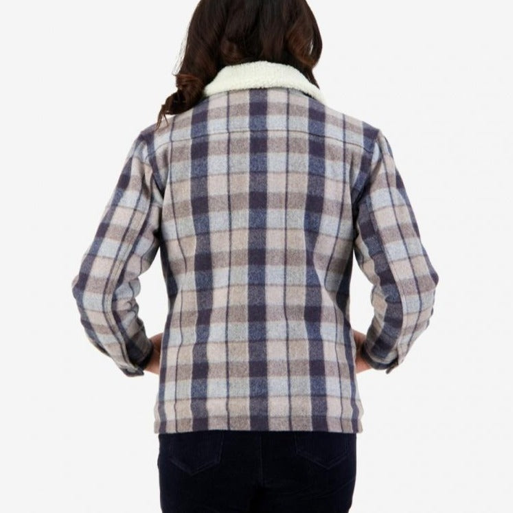 A person with long hair is shown from behind wearing the Swanndri Kaituna Women's Sherpa Lined Jacket, a stylish plaid jacket in shades of blue, gray, and beige featuring a white sherpa collar. Made from a wool cotton blend, they pair it with dark-colored pants. The background is plain and light-colored.
