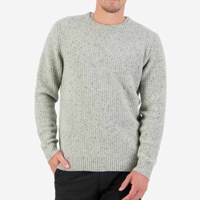 A person is standing against a white background, dressed in the Swanndri Sentry Hill Knit Crew, a light gray casual knitwear sweater with a crew neckline, paired with dark pants. One hand is in their pocket while the other rests by their side.