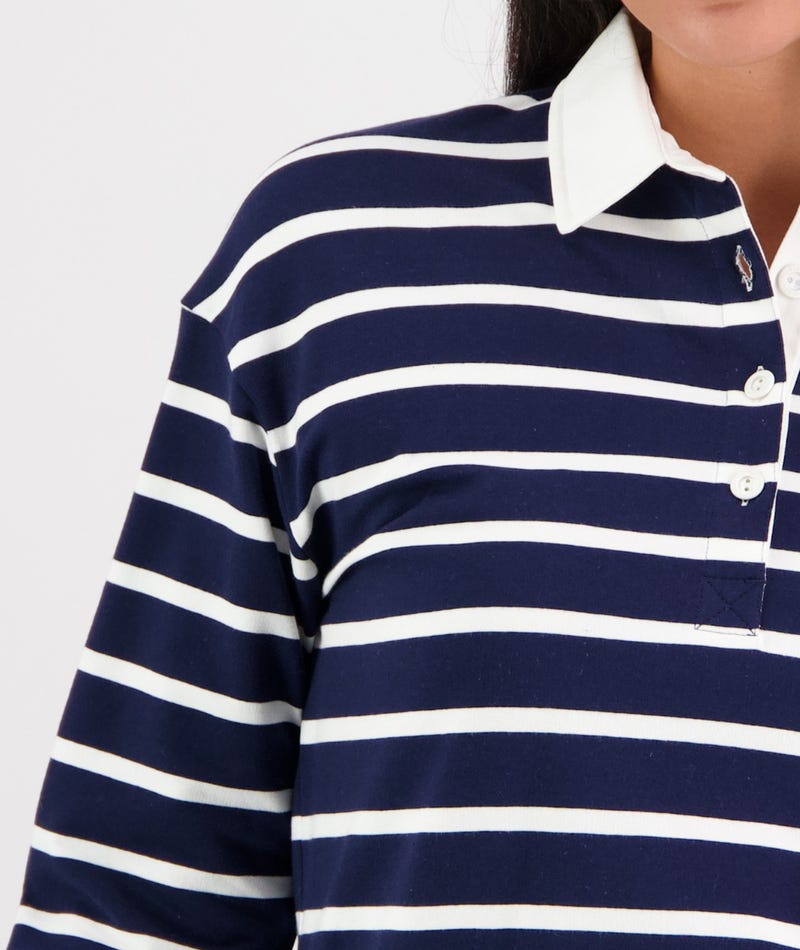 A person wearing the Swanndri Carlaw Rugby shirt, which is navy blue and white striped with long sleeves and a white collar. The shirt, reminiscent of a casual rugby style, features buttons down the front and a small embroidered logo near the collar. The photo is cropped to show only the upper torso and part of the neck.