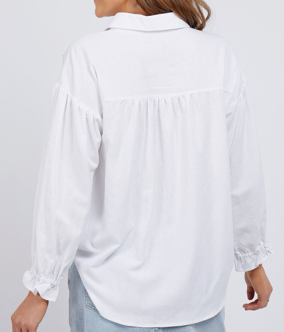 A person with long hair is facing away from the camera, wearing a loose-fitting Foxwood Mia Shirt in white, featuring ruffled cuffs and paired with light blue jeans. The slightly gathered back yoke of the viscose linen blend shirt adds a touch of elegance. The background is plain and white.