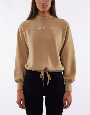 A person wearing a beige Silent Theory Sleek Crew jumper with "Silent Theory Co." printed on the chest and black pants stands against a plain white background. The jumper features long sleeves, a tie waist, and a relaxed fit. The person's face is not visible.