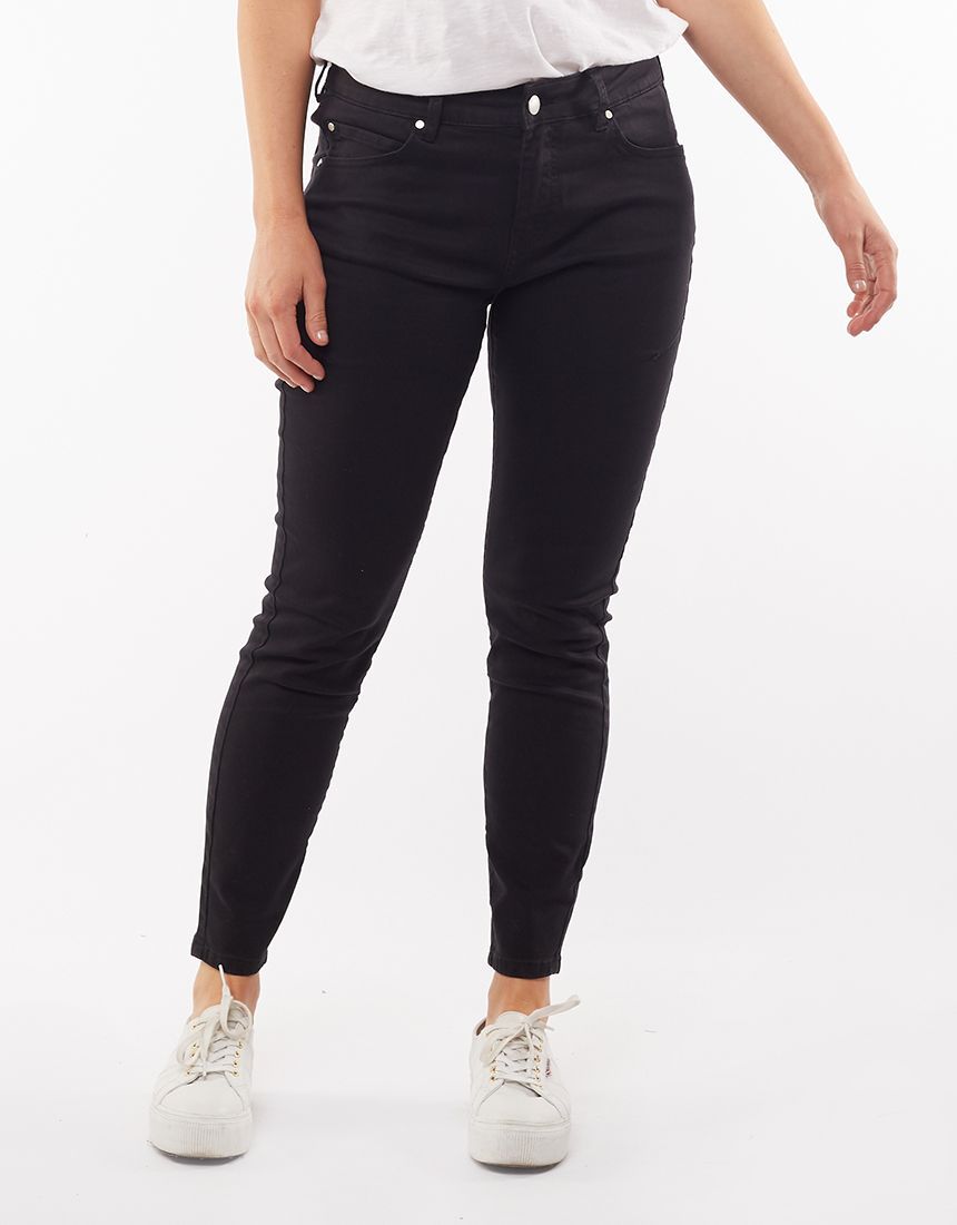 A person stands against a plain white background, wearing Foxwood City Jean in black with a classic five-pocket design, paired with a white t-shirt and white sneakers. Their hands are relaxed by their sides.