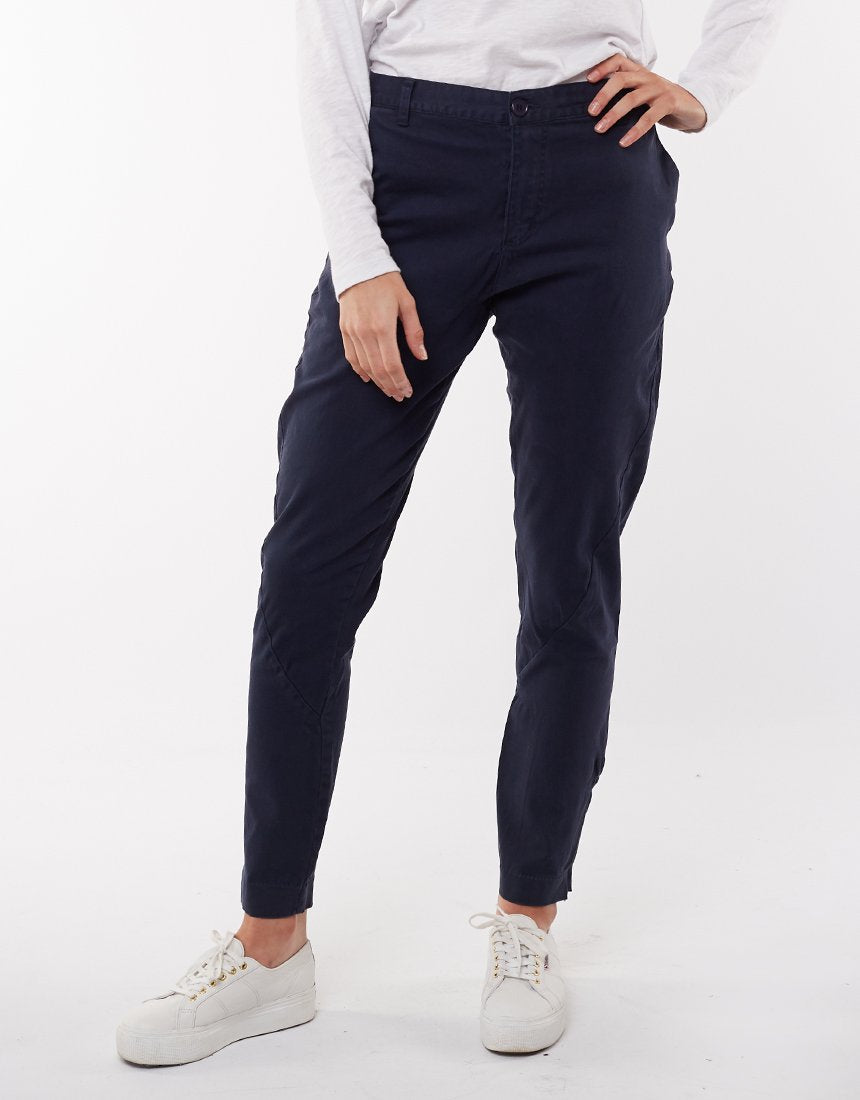 A person in navy blue Foxwood Liberty Pants and a white long-sleeve shirt is standing with their right hand placed on their hip. They are also wearing white sneakers. The comfortable pants have a relaxed fit with a tapered leg, perfect for casual wear. The background is plain white.