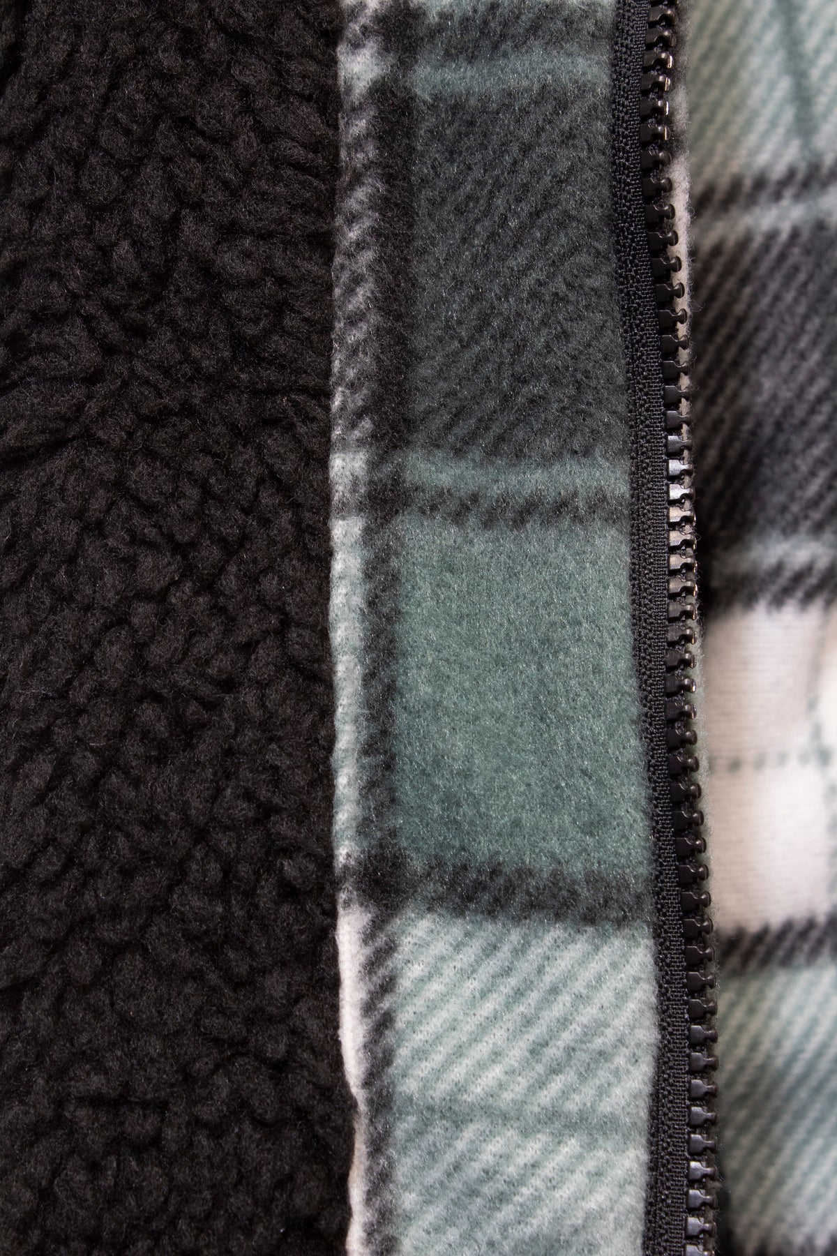 Close-up of the Pilgrim Sherpa Zip Jacket fabric displaying two textures: a black sherpa fleece lining on the left and a green plaid pattern on the right, divided by a black zipper. The plaid showcases shades of green, white, and gray in durable polyester.
